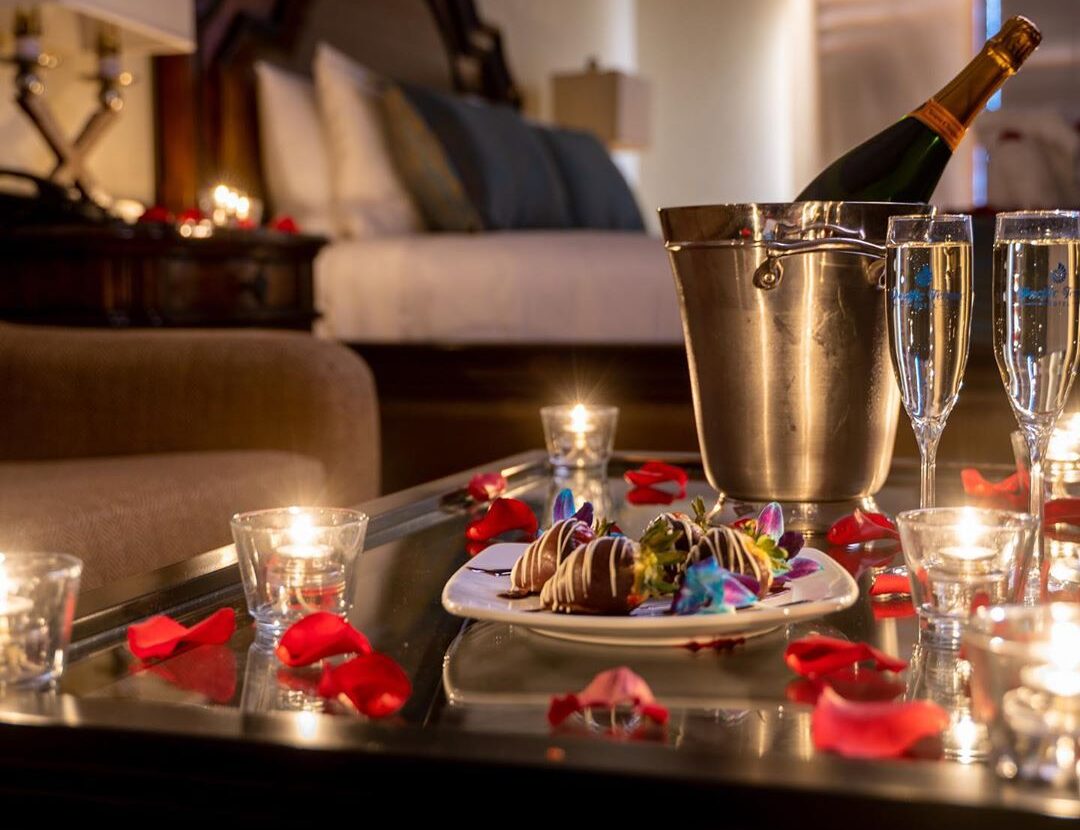 bottle of champagne and chocolate covered strawberries on table in front of bed